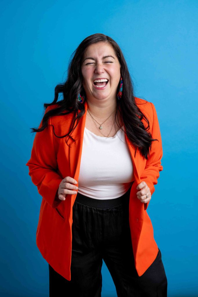 Woman with long dark hair faces the camera, smiling. She wears an orange blazer. Background is blue.